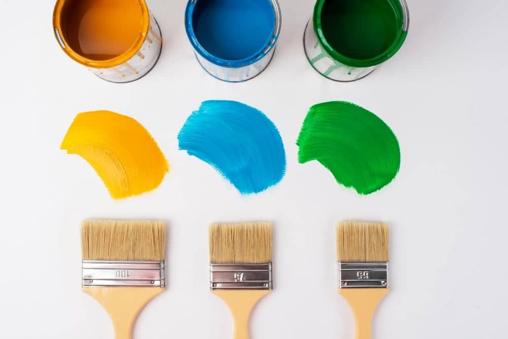 Three paint brushes and cans of yellow, blue, and green paint arranged on a white surface, showing how different colors can transform and personalize a DIY TV stand.