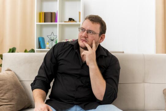 thoughtful adult slavic man optical glasses sits armchair putting hand chin inside living room 1 scaled - best couches for heavy person, couches