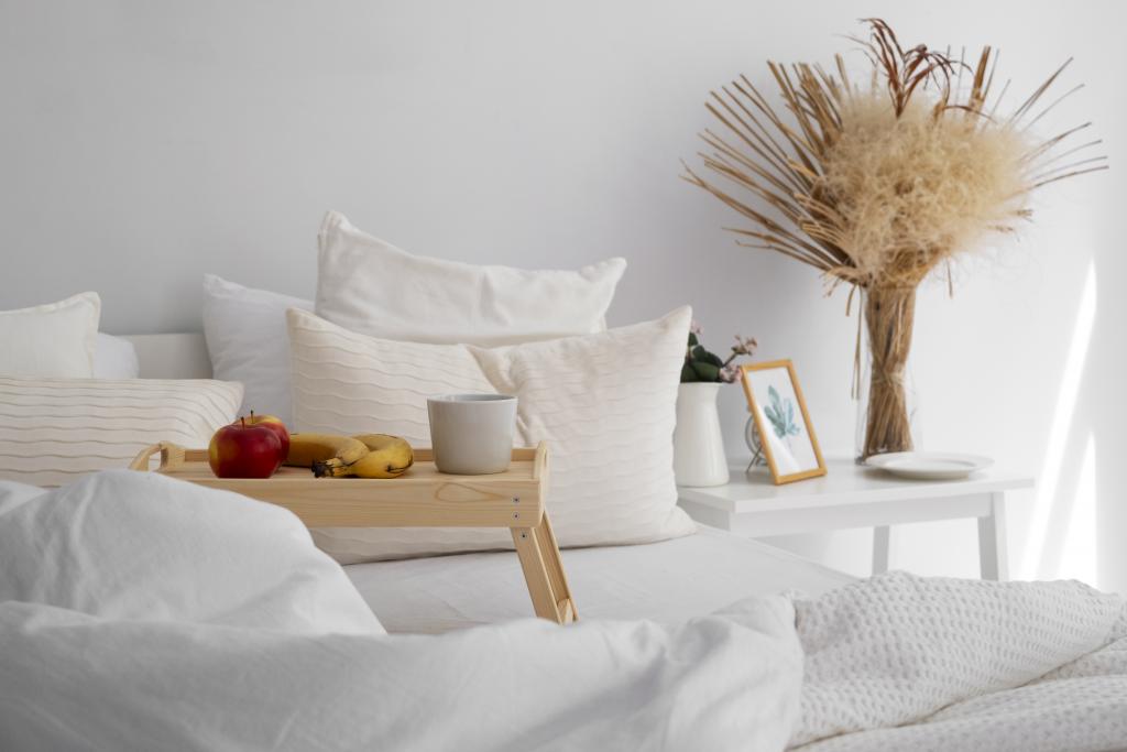 Cozy minimalist white bed featuring a breakfast tray, a small table, a small potted plant, and framed botanical print.